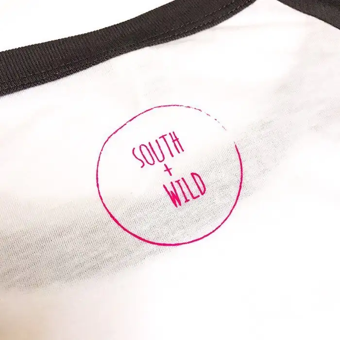 South + Wild logo printed in pink inside a circle on the back nape of a white shirt